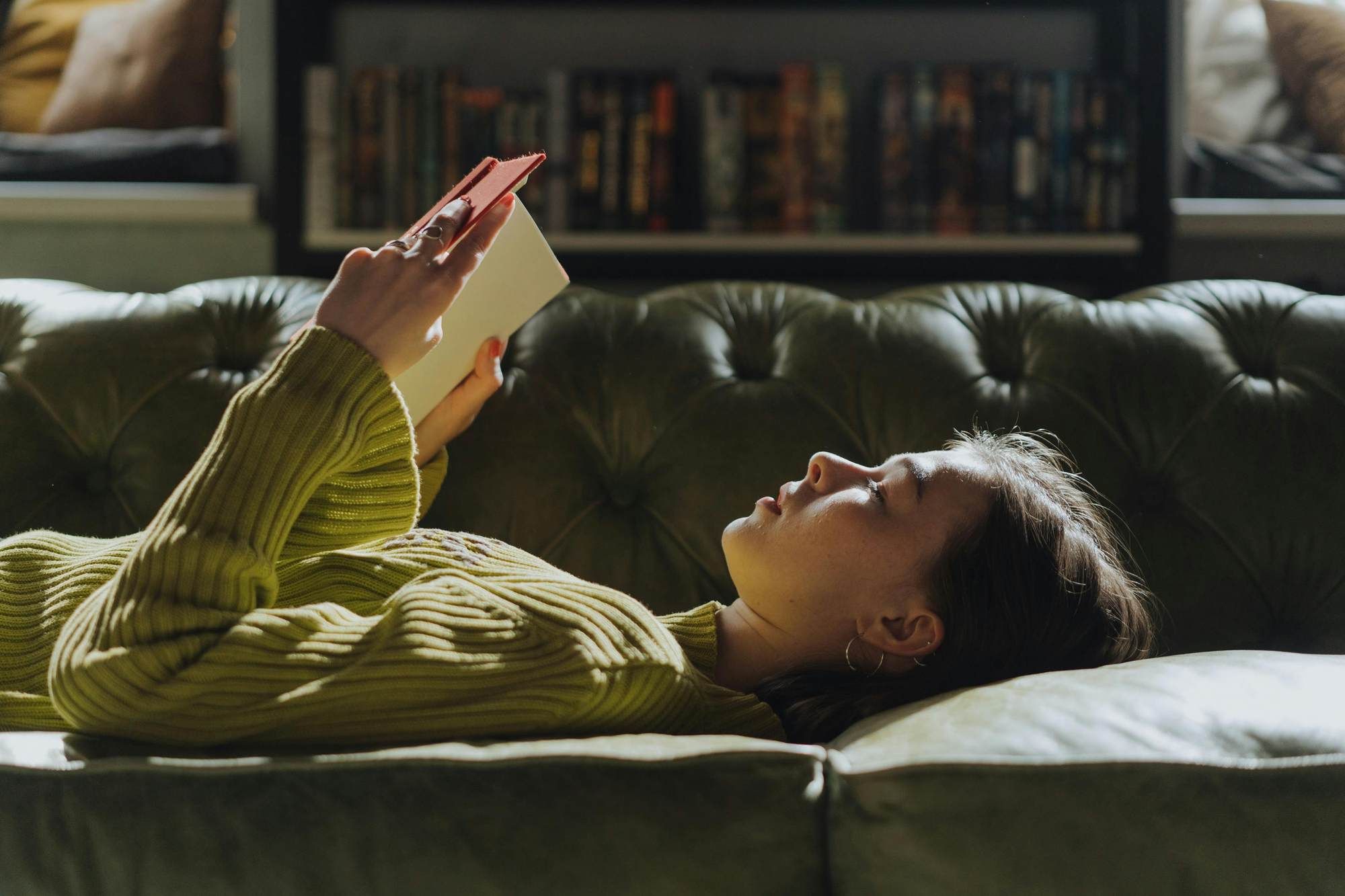 A person in a green sweater lies on a green sofa, holding and reading a book, with bookshelves in the background.