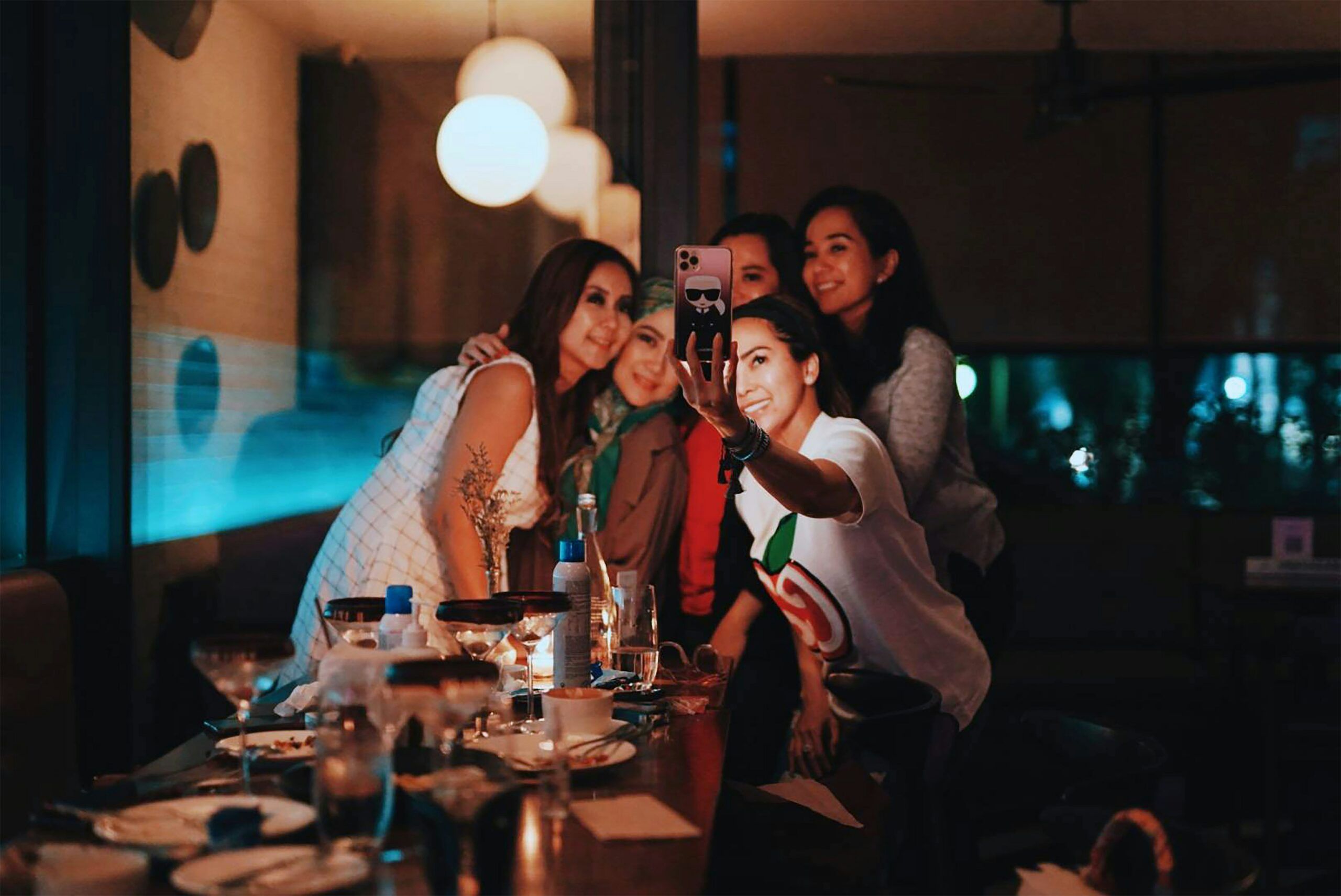 Five women are posing for a group selfie at a restaurant table with food and drinks.